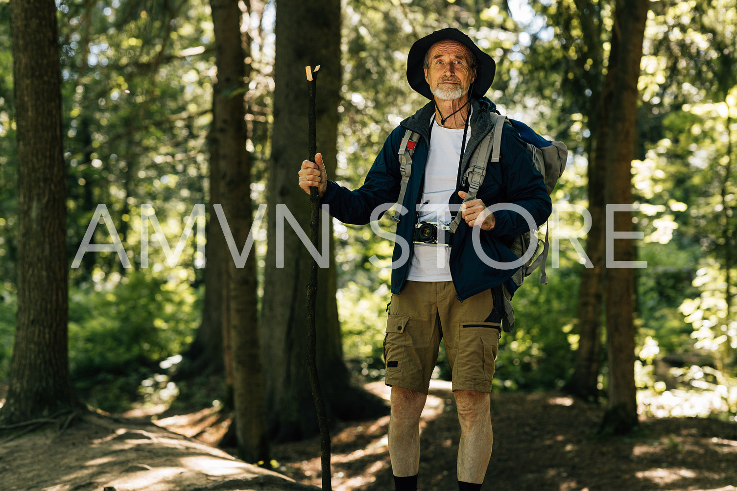 Senior male with a wooden stick standing in a forest with a backpack. Portrait of a mature man standing in forest.