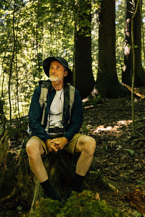 Full-length senior male with a hat, backpack, and hiking clothes sitting in the forest and looking away