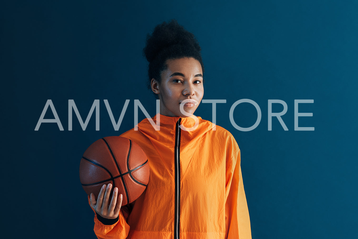 Portrait of young female basketball player against blue backdrop. Woman in orange sportswear holding a basketball looking at camera.