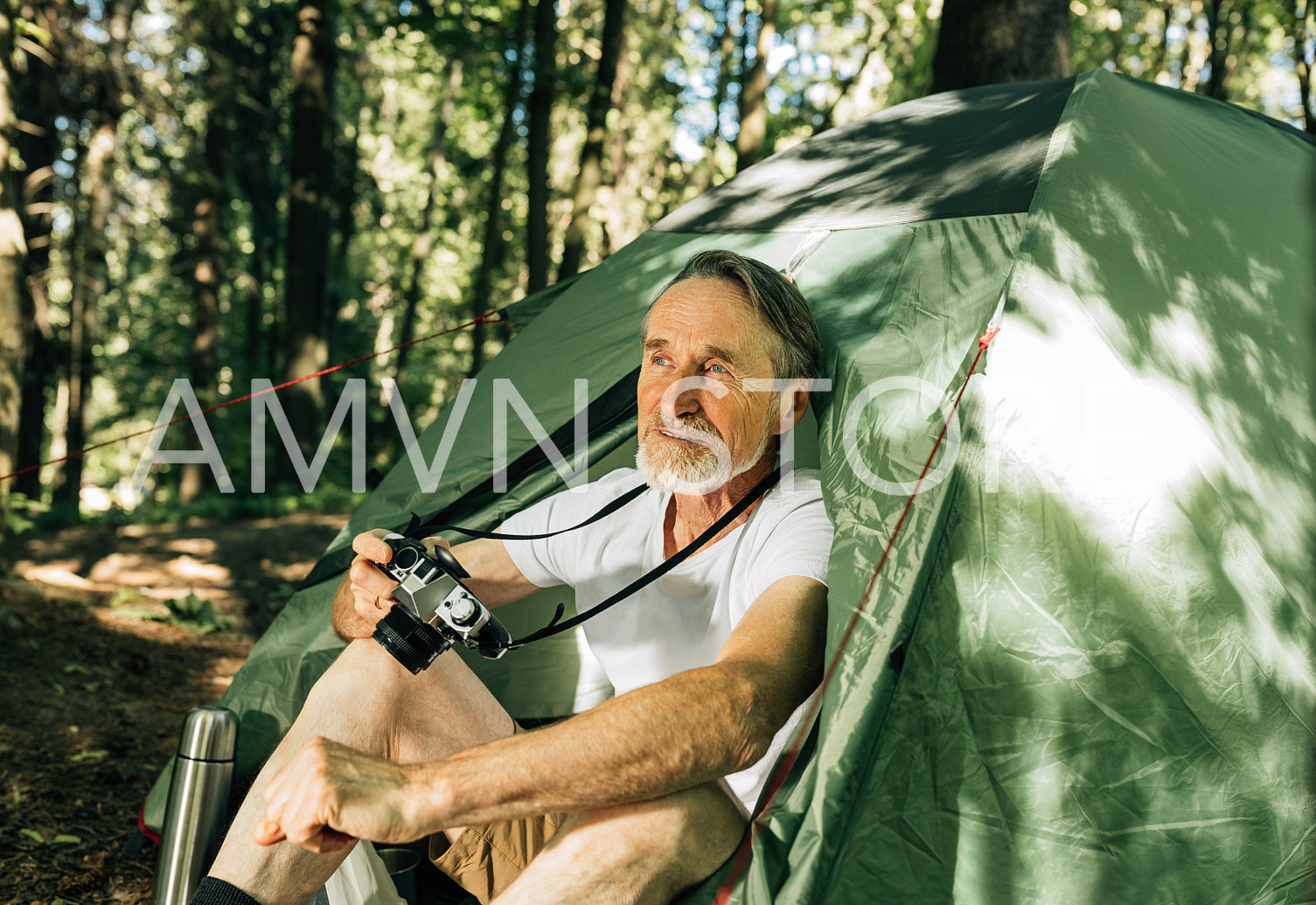 Senior tourist looking away while sitting in tent. Mature male with a film camera enjoying his forest camping.