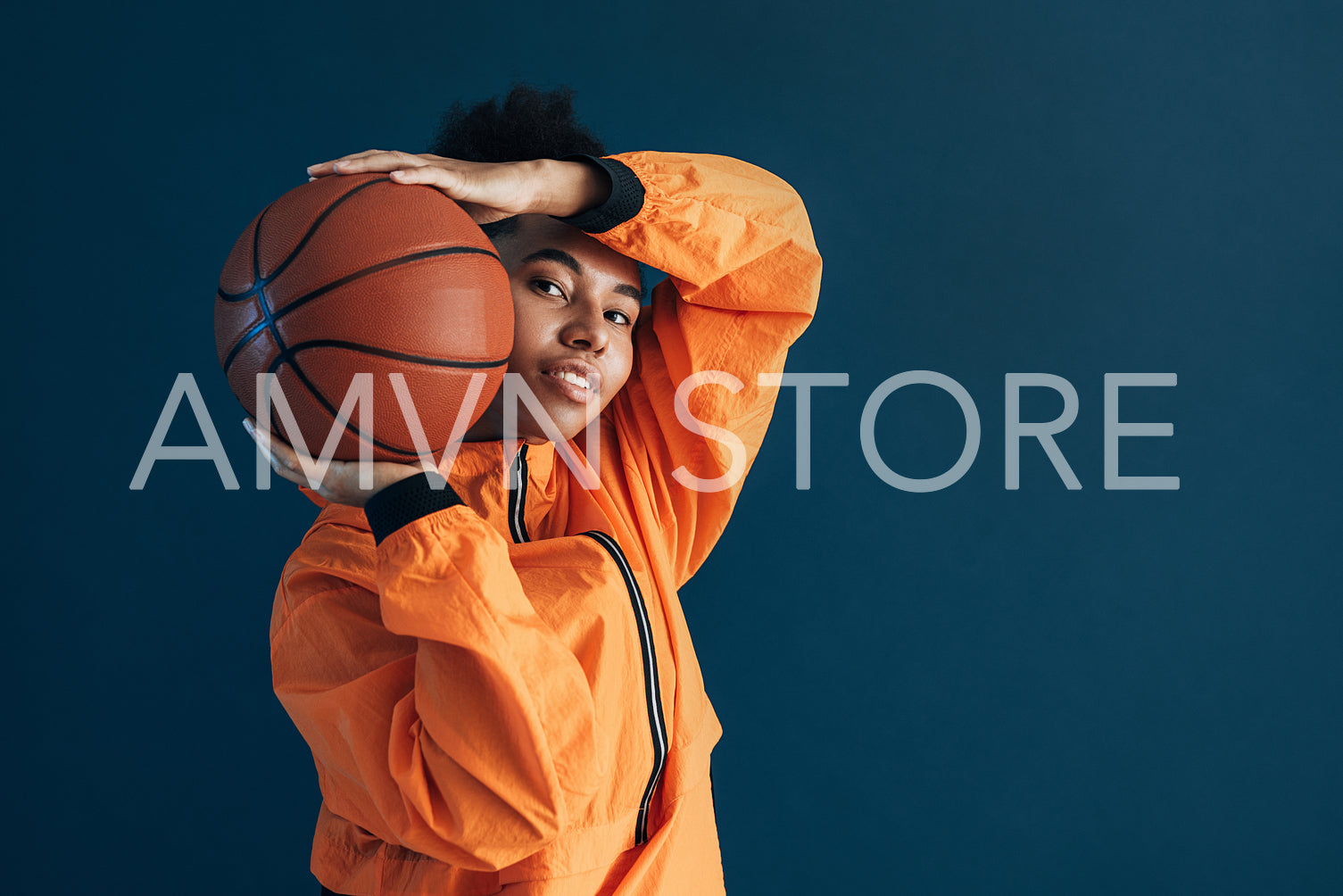 Portrait of a young basketball player posing with ball against blue backdrop and looking at camera