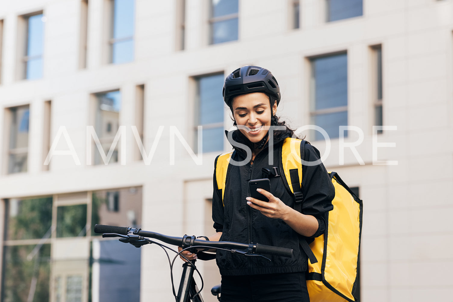 Female courier in cycling helmet with bicycle in the city. Smiling woman with thermal backpack looking at smartphone.