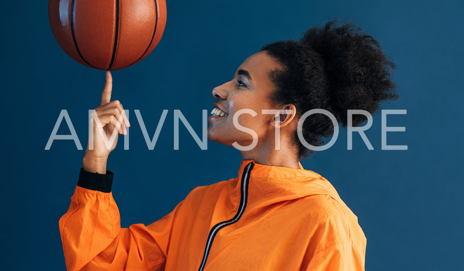 Side view of smiling woman spinning basketball on her finger while posing in studio against blue backdrop