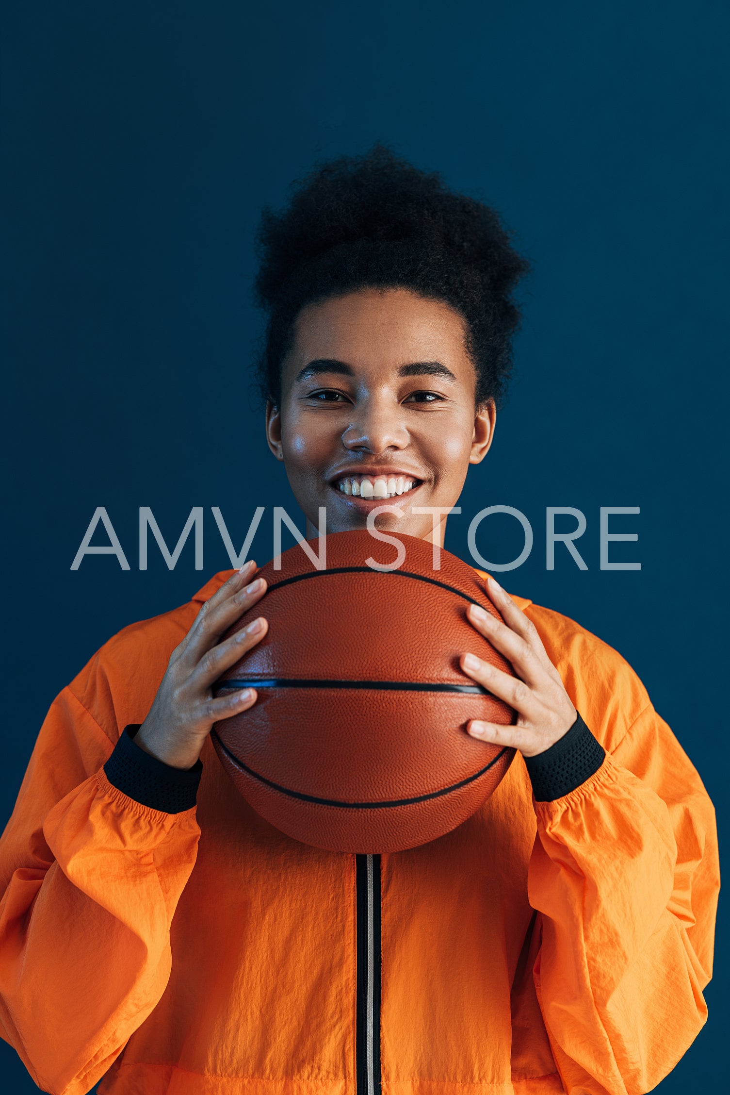 Portrait of a young smiling basketball player. Cheerful female in orange sportswear holding basketball over blue backdrop.