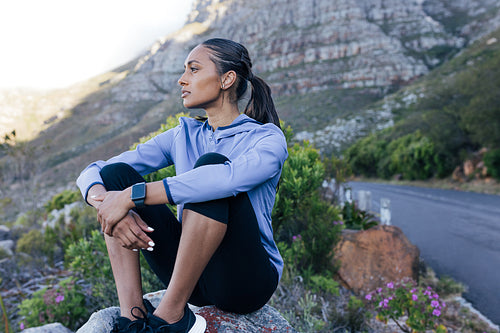 Young runner sitting outdoors on a rock in a natural park. Female in fitness attire enjoy the view while taking a break during outdoors workout.