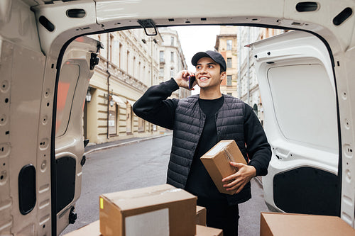 Smiling courier in uniform holding box and making call to a cust