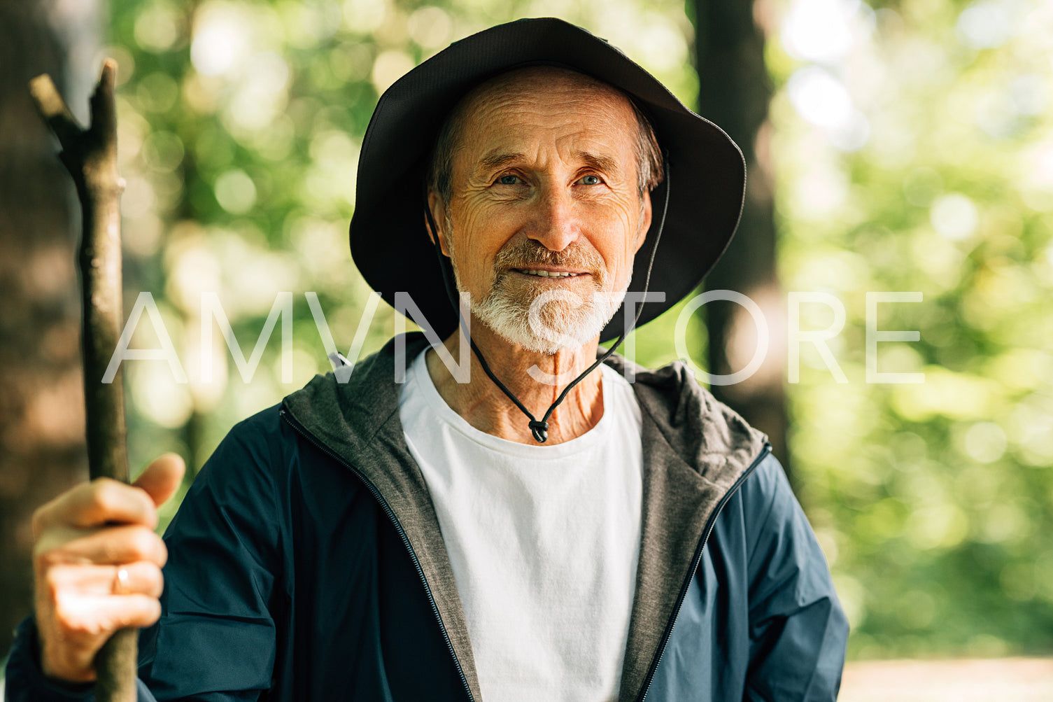 Portrait of a senior male wearing a hat and holding a stick. Mature man looking at the camera during his forest walk.