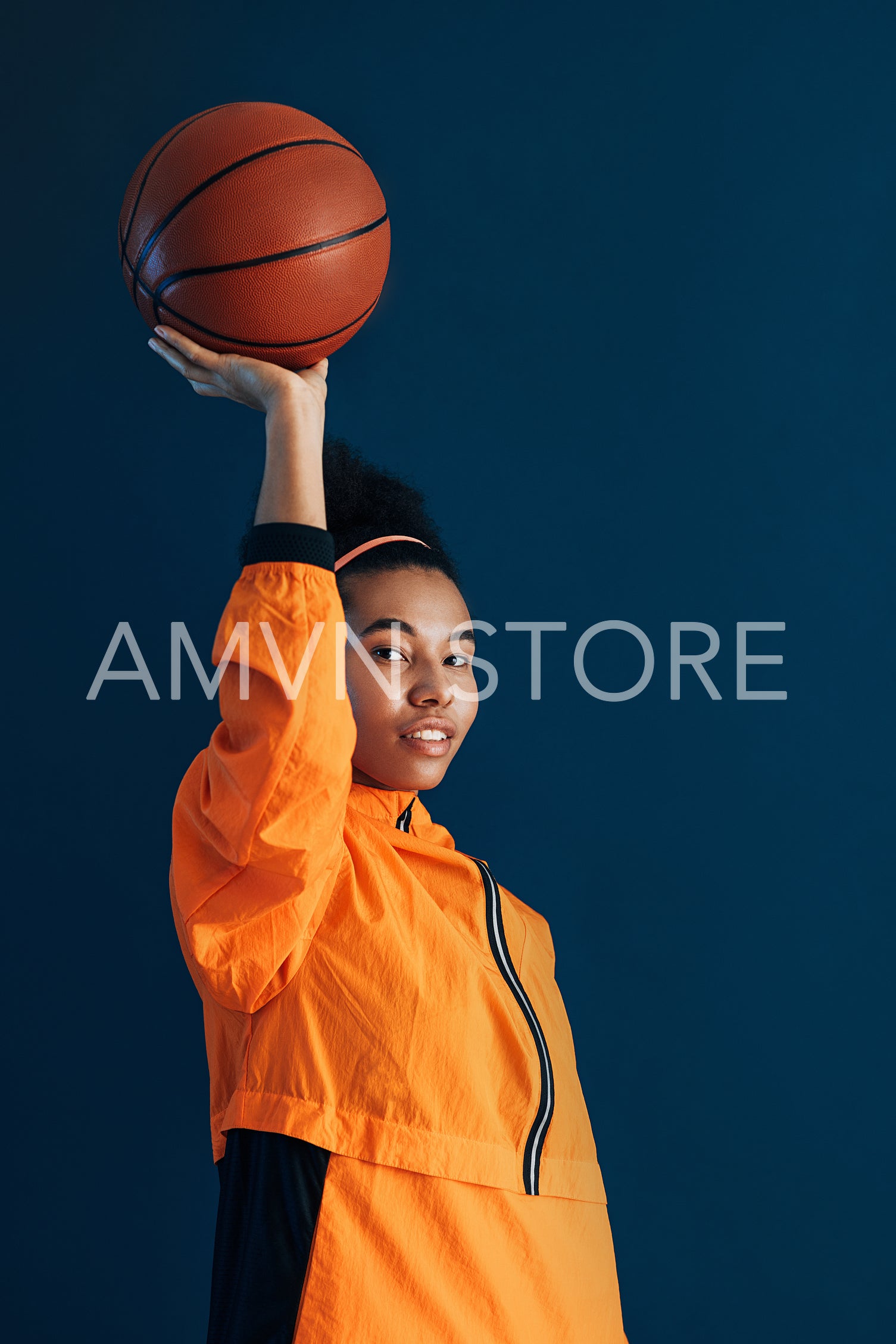 Young female basketball player holding a basketball on an outstretched arm. Studio portrait of a woman basketball player in orange sportswear.