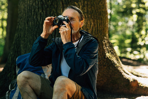 Senior male sitting at tree and taking photos on film camera. Mature man taking a break during forest walk.