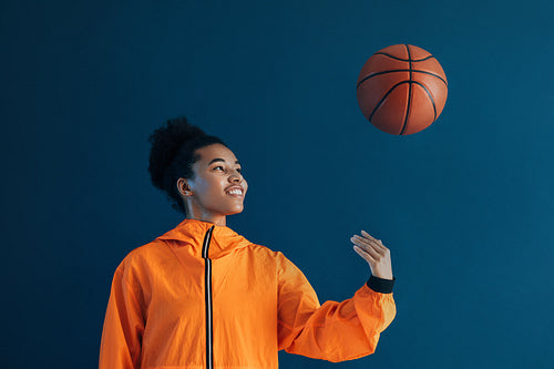 Female basketball player tossing a basketball over a blue background. Young woman in orange sportswear tossing basketball up in studio.