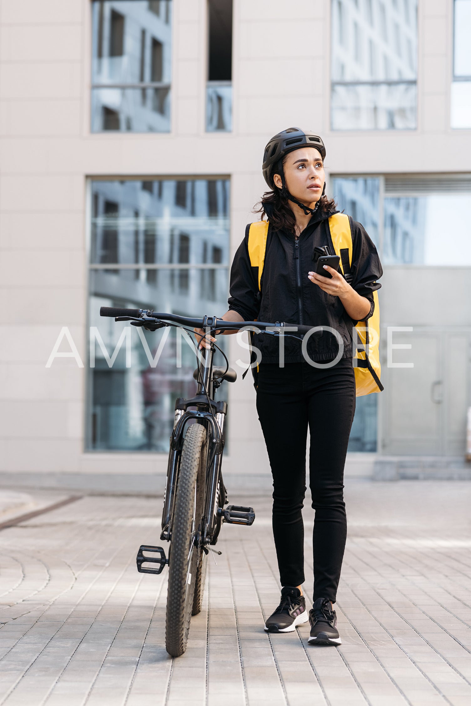 Woman courier walking on a city street with bicycle, holding mobile phone looking away