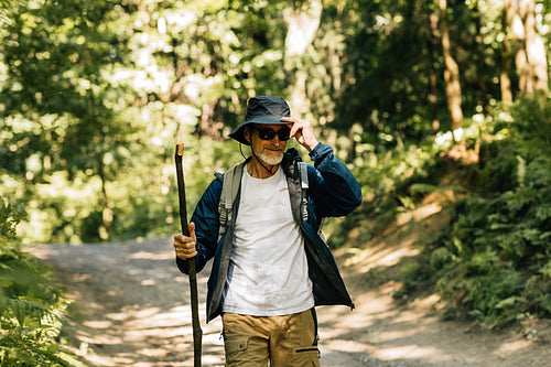 Senior male with a wooden stick wearing hat and sunglasses while trekking in forest