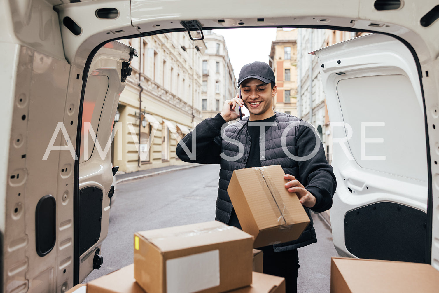 Smiling courier in uniform looking at box and making call to a c