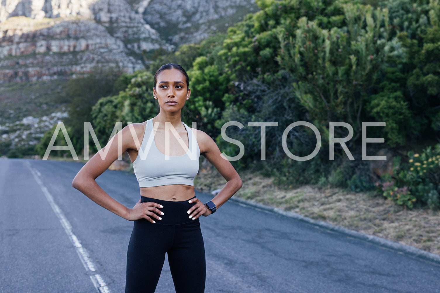 Confident female runner standing on an abandoned road and looking away. Slim woman in fitness attire with hands on hips relaxing during a run in the park.