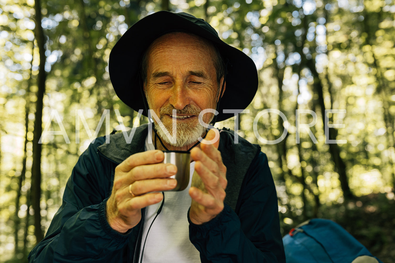 Cheerful male in a hat sitting is the forest drinking hot coffee from a metallic cup