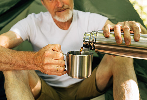 Close-up of a senior male pouring coffee from a thermos to a metallic cup while sitting tourist tent