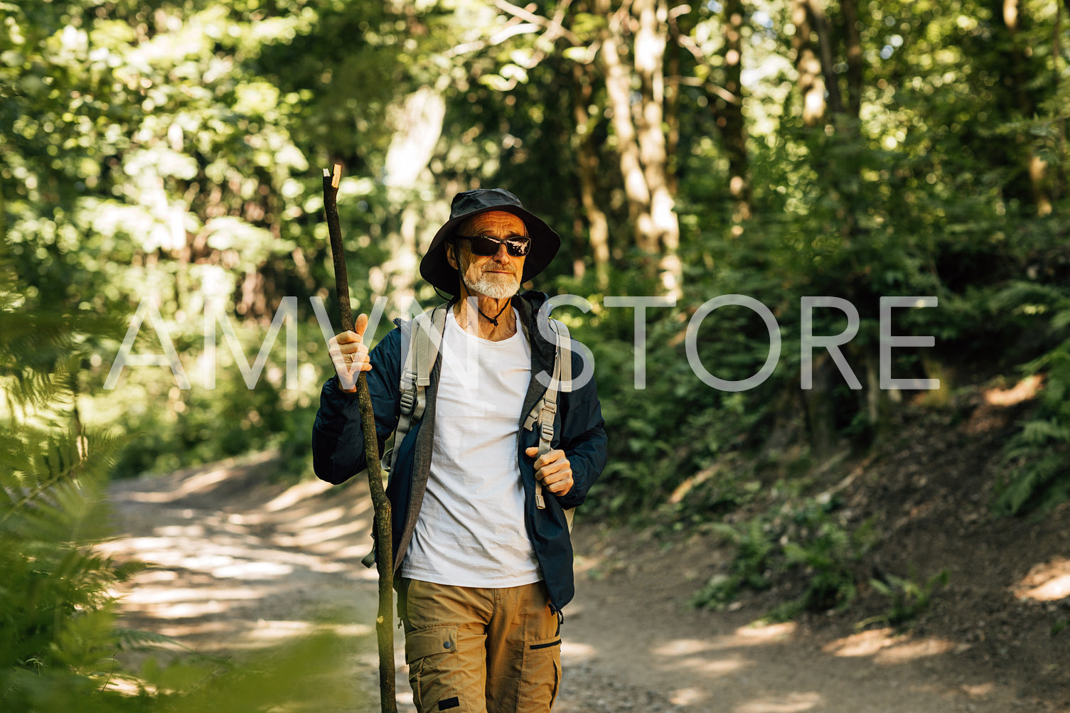 Senior man wearing glasses, hat and hiking clothes walking in a forest with a wooden stick