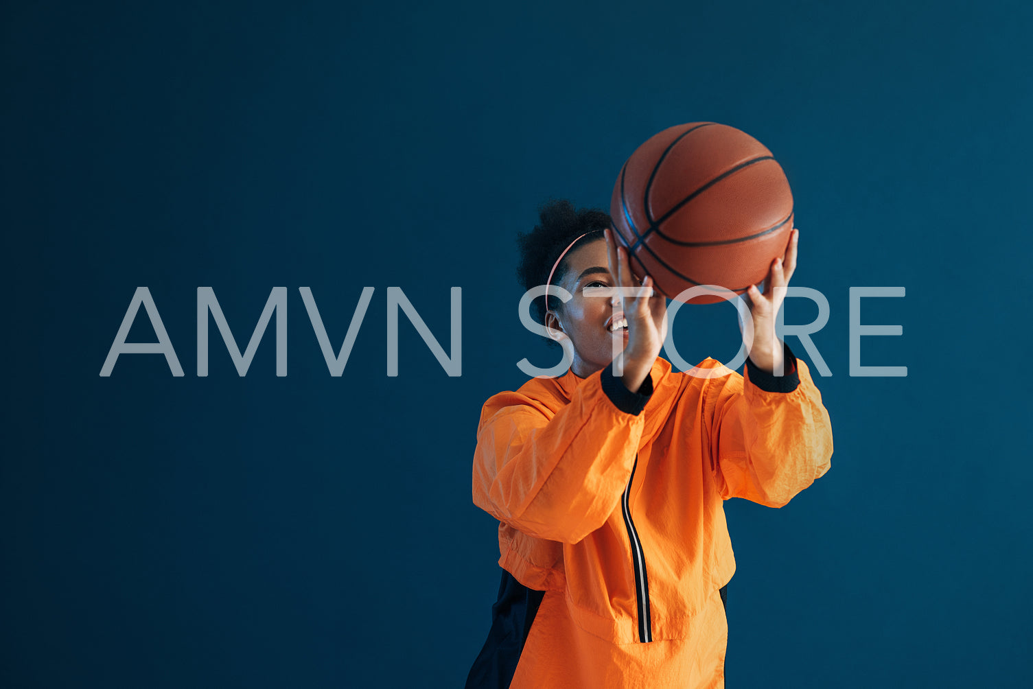 Smiling female basketball player throws basketball over blue backdrop in studio. Young woman in orange fitness wear practicing with basketball.