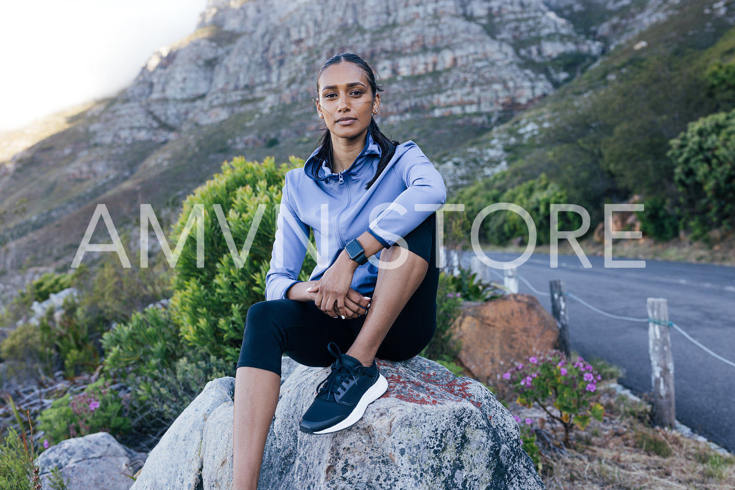 Young slim female sitting on rock relaxing after a trail run. Woman in sportswear looking at camera while sitting in natural park.