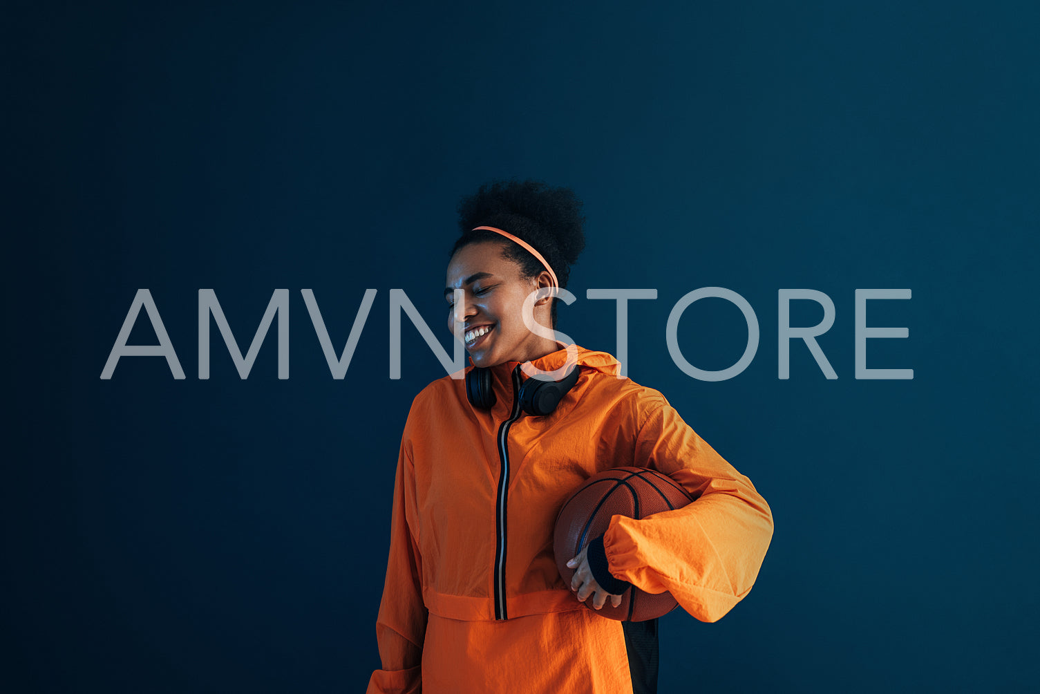 Cheerful female in orange sports attire over blue backdrop. Young smiling professional basketball player in the studio.