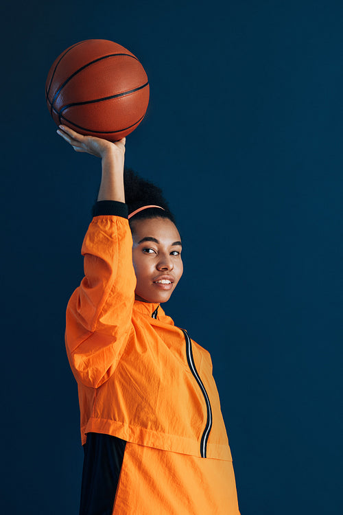 Young female basketball player holding a basketball on an outstretched arm. Studio portrait of a woman basketball player in orange sportswear.