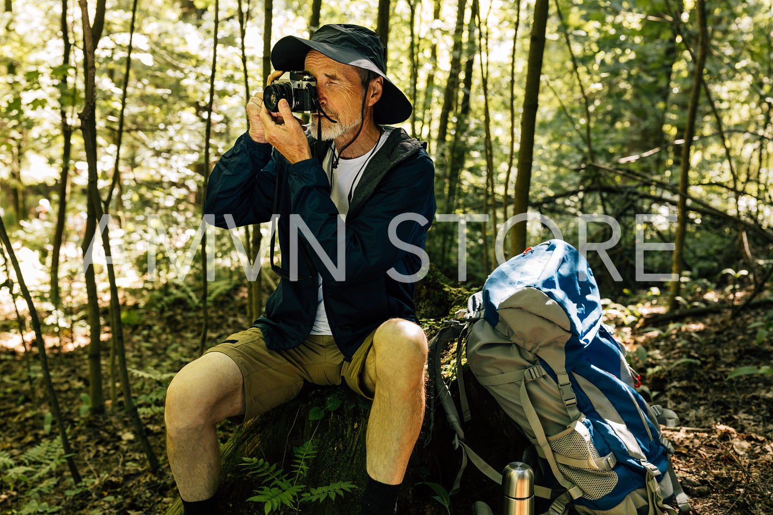 Senior man sitting on tree stump and making photographs on film camera. Male with retro camera and backpack making pictures.