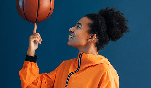 Side view of smiling woman spinning basketball on her finger while posing in studio against blue backdrop