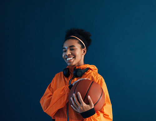 Happy sportswoman with closed eyes holding basketball while standing at blue backdrop in studio. Cheerful female posing with basketball in studio.