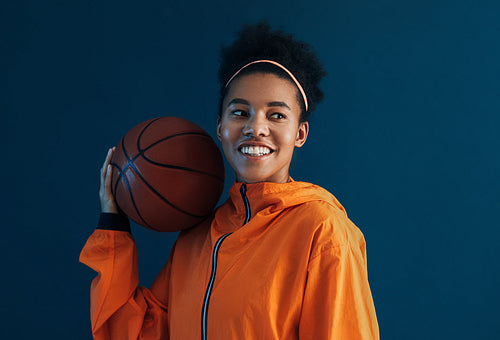 Portrait of a young and cheerful female basketball player. Woman in orange fitness attire holding a basketball on a shoulder and looking away over a blue background.