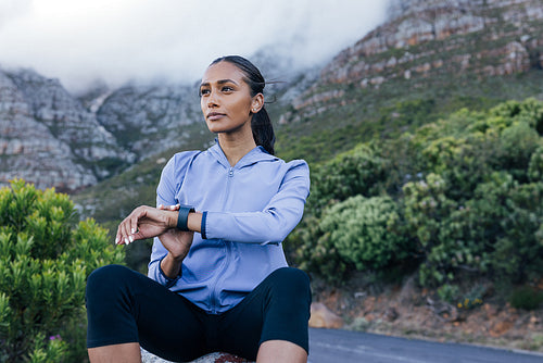 Slim woman in fitness attire sitting on the rock. Female using smartwatches looking into the distance.