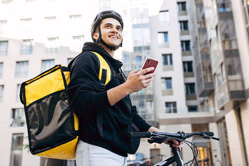 Portrait of delivery man holding a mobile phone, wearing thermal backpack standing against apartment building