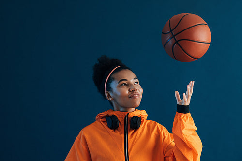 Cheerful female in orange fitness attire throw a basketball over blue background. Smiling sportswoman looking at flying basketball.