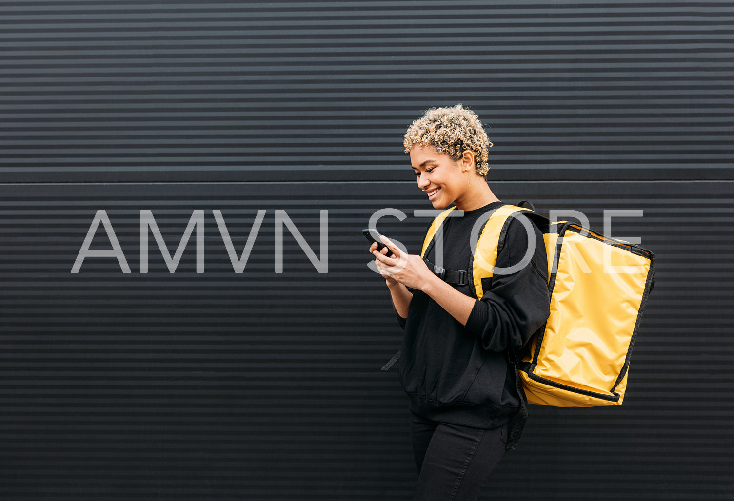 Smiling courier girl with food delivery backpack holding a smartphone while standing at black wall outdoors