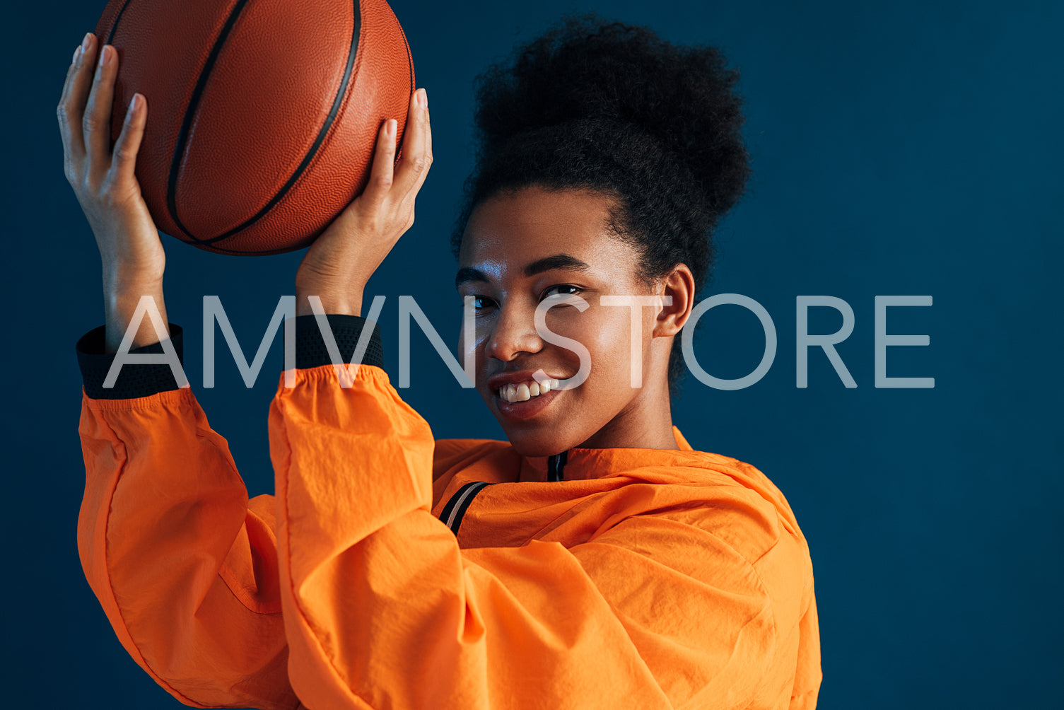 Close-up portrait of a young smiling basketball player looking at the camera over a blue backdrop. Female with curly hair with basketball wearing orange sportswear.