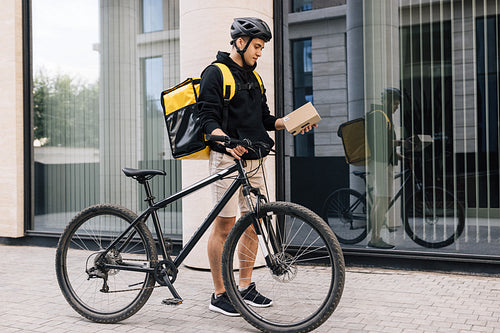 Young man working for delivery company standing near an office building looking on a cardboard box