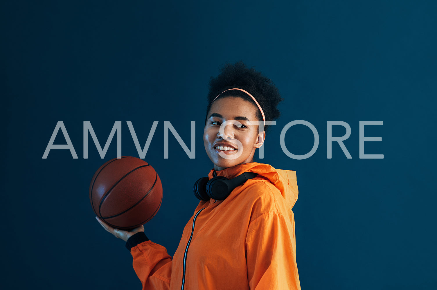 Young smiling female holding basketball on one hand and looking away while standing over blue backdrop. Positive sportswoman in orange fitness wear over blue background.