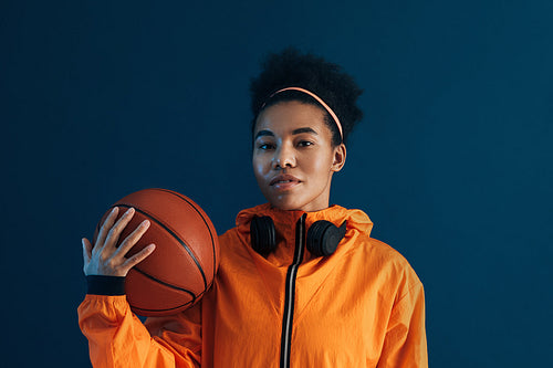 Portrait of confident female basketball player looking at camera in studio. Young woman with basketball posing over blue backdrop.