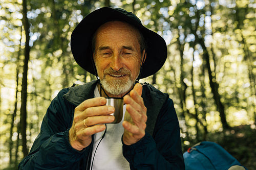 Cheerful male in a hat sitting is the forest drinking hot coffee from a metallic cup