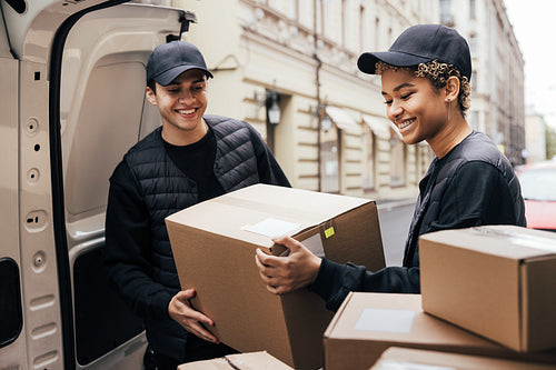 Two smiling couriers unloading cardboard boxes from van in the c