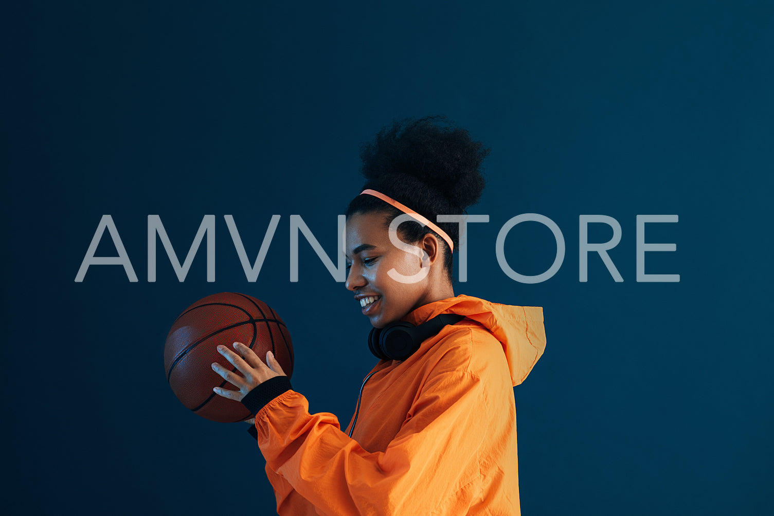 Smiling sportswoman in orange clothes holding a basketball. Side view of a young female with headphones holding basketball over a blue backdrop.