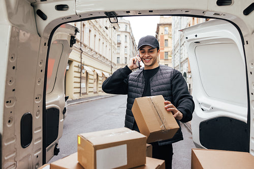 Smiling courier in uniform looking at box and making call to a c