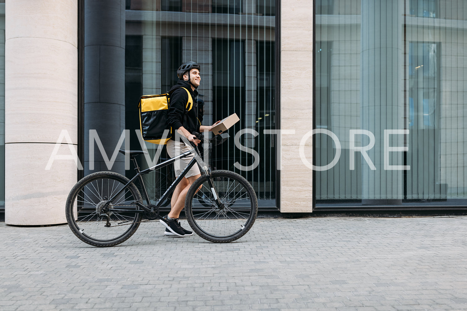 Side view of a male courier walking with a bicycle holding a package. Delivery man with thermal backpack at an office building.