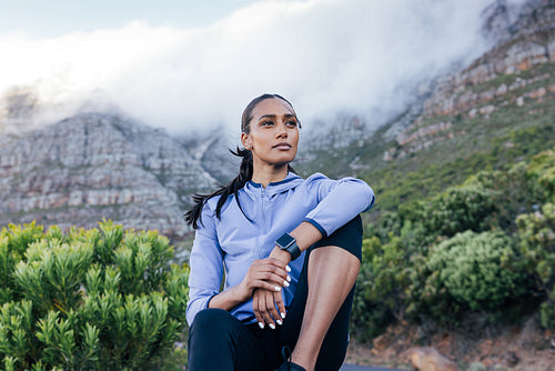 Confident slim female sitting against the stunning view in a natural park. Young woman in sportswear relaxing outdoors and looking away.