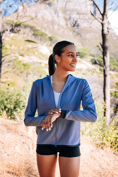 Side view of cheerful woman athlete looking away. Smiling female checking smartwatch outdoors.