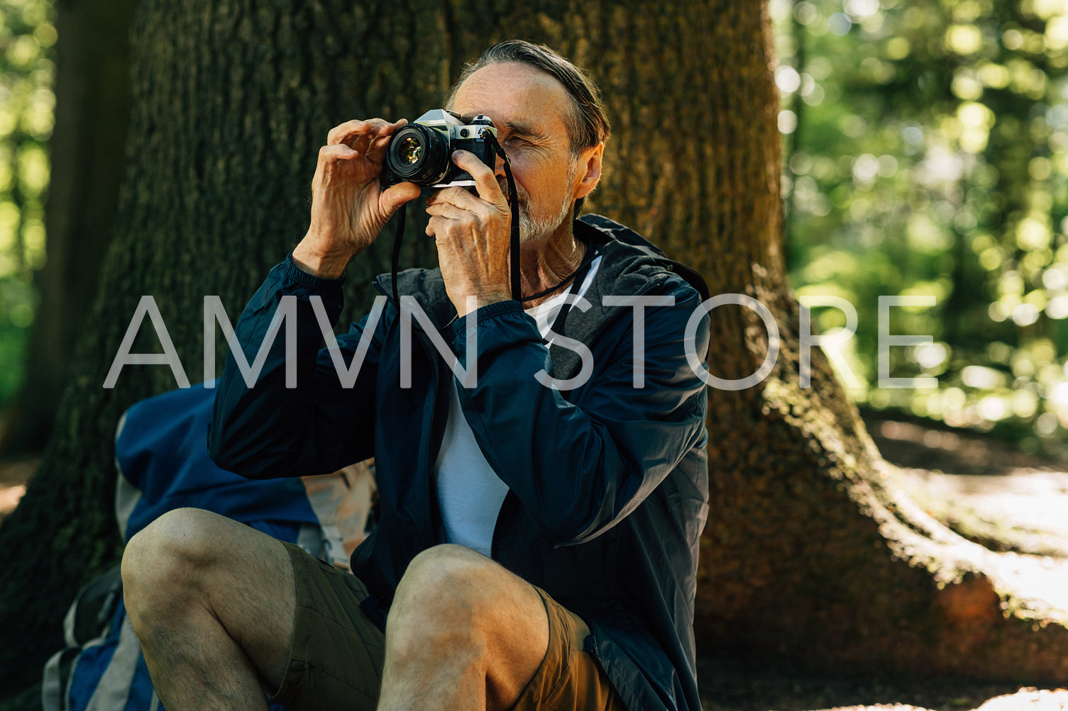 Senior male sitting at tree and taking photos on film camera. Mature man taking a break during forest walk.