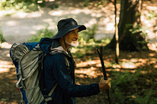 Cheerful senior male with a wooden stick and tourist backpack walking in a forest