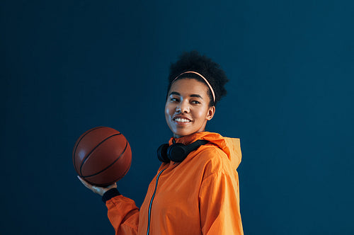 Young smiling female holding basketball on one hand and looking away while standing over blue backdrop. Positive sportswoman in orange fitness wear over blue background.