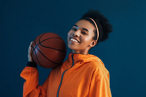 Cheerful basketball player posing with basketball in the studio over blue backdrop. Smiling female in orange sportswear.