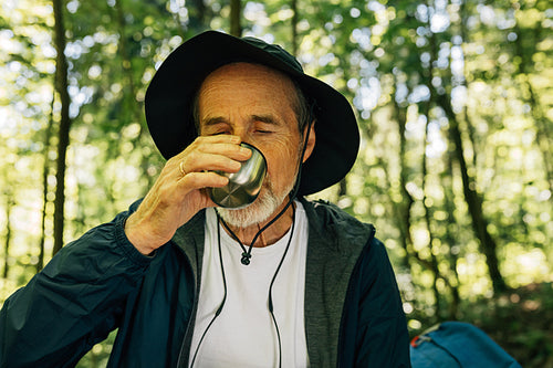 Senior male in hat drinking during his outdoor walk in forest