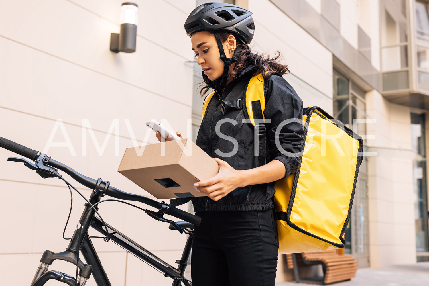 Side view of female messenger wearing cycling helmet and backpack looking on mobile phone while holding a parcel